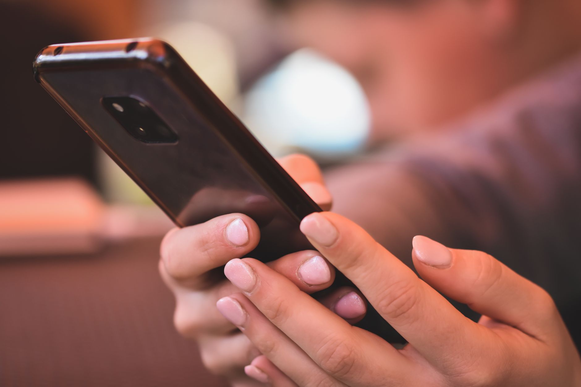 Deux mains tiennent un smartphone sombre, dont on voit le dos et l’objectif, devant un arrière-plan flou d’intérieur.