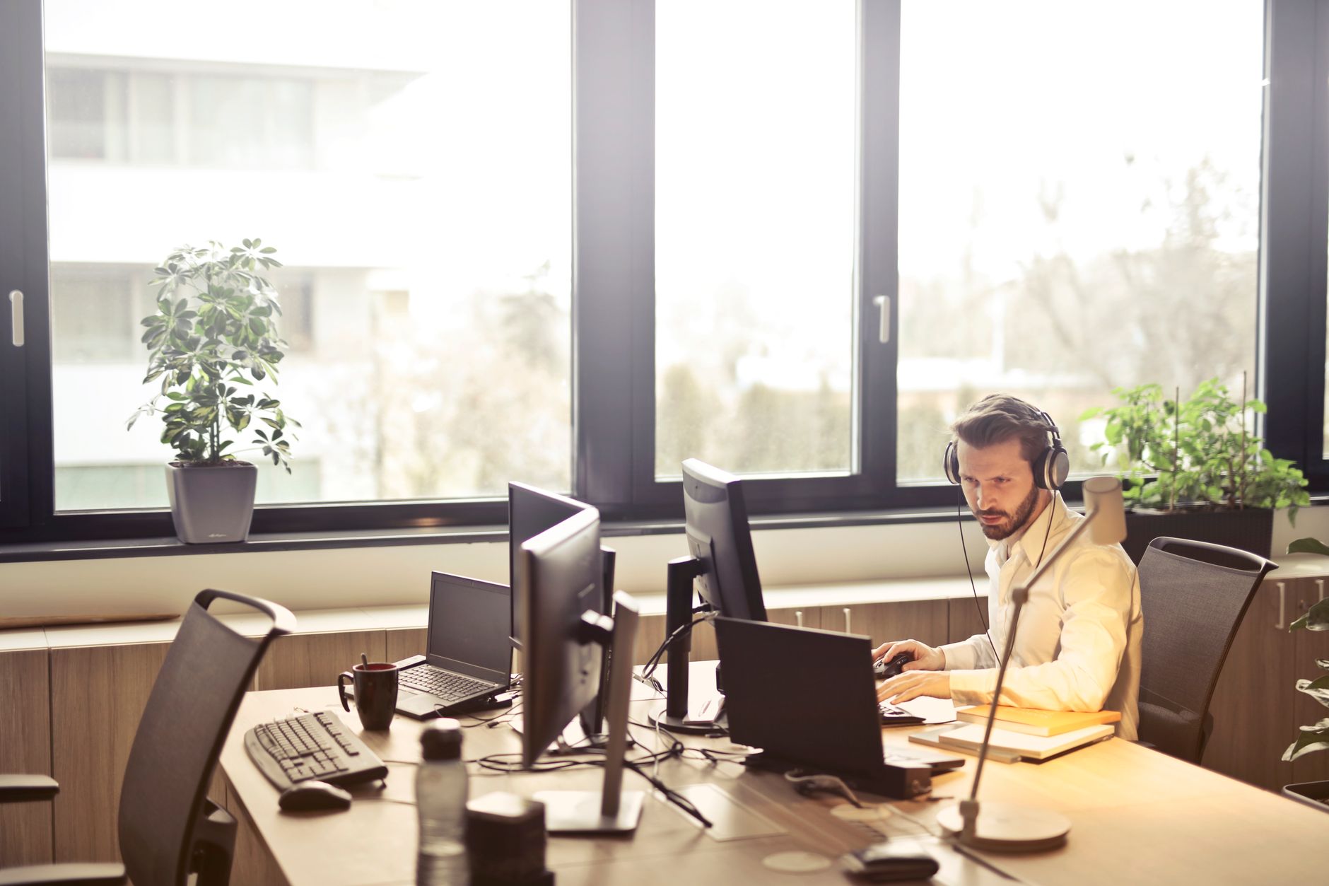 Un homme avec un casque audio travaille à un bureau équipé de plusieurs écrans et ordinateurs portables, devant de grandes fenêtres.